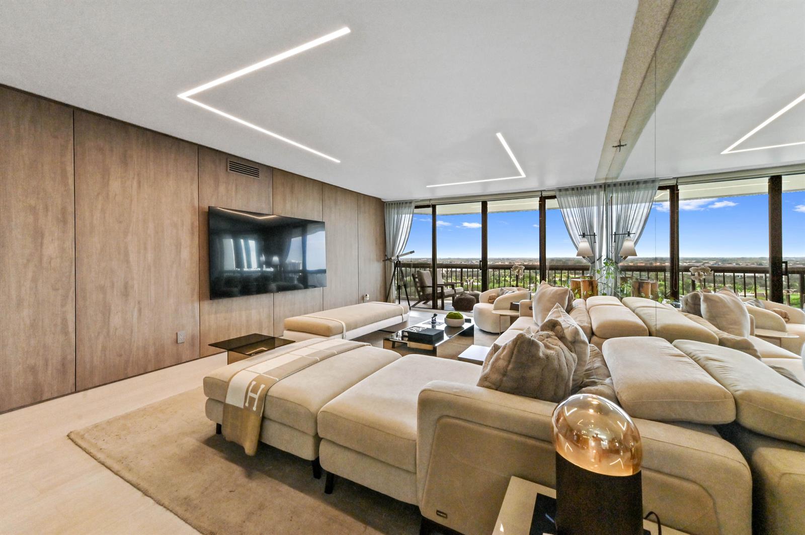 A modern living room features wood-paneled walls, a large sectional sofa, and floor-to-ceiling windows — 1900 Consulate Place # 1205, West Palm Beach FL 33401, WEST PALM BEACH, FL