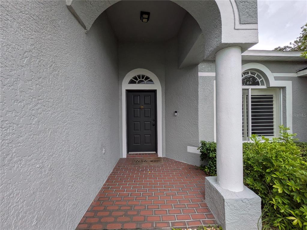 A gray front door with a transom window sits under an arched entryway with brick flooring — 16120 MUIRFIELD DRIVE, ODESSA, FL