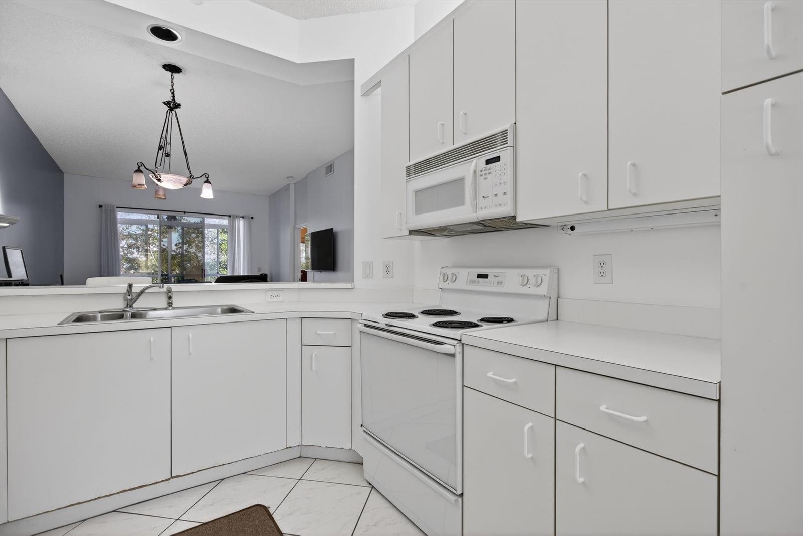A white kitchen features a stove, microwave, sink, and tile flooring, opening to a living area — 9575 Weldon Circle Apt C413, Tamarac FL 33321, TAMARAC, FL