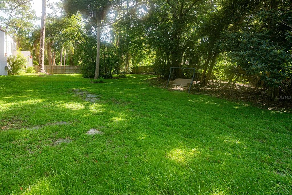 A grassy backyard features mature trees, a wooden fence, and a metal swing set — VALENCIA STREET, SANFORD, FL
