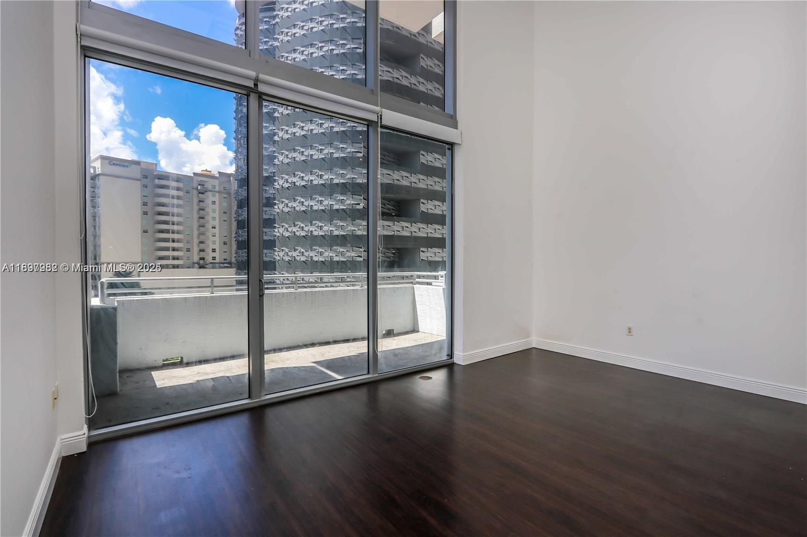 An empty room with dark wood flooring, tall windows, and a balcony overlooking city buildings — 1050 Brickell Ave # 414, Miami FL 33131, MIAMI, FL