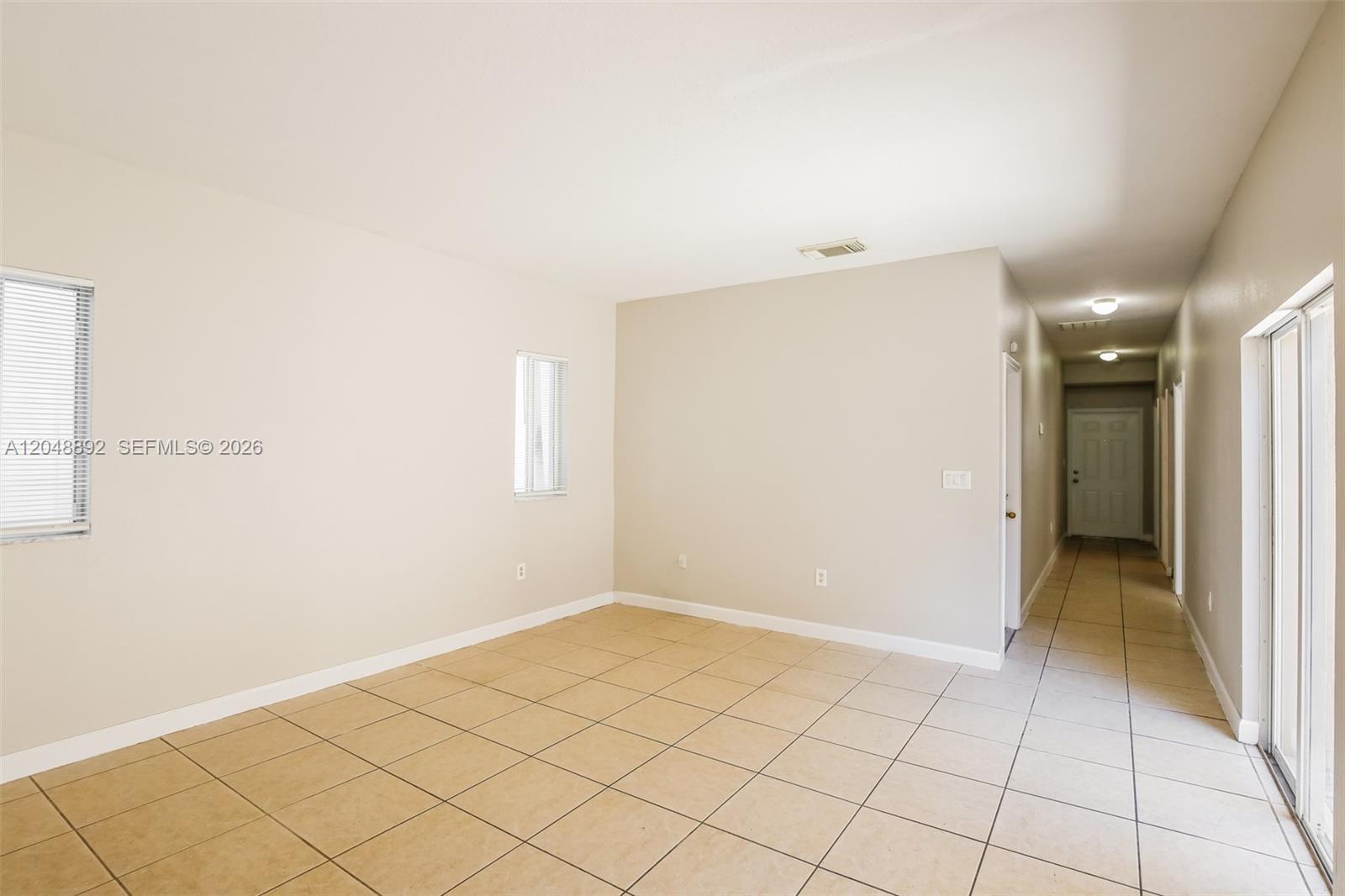 An empty room with beige tile flooring, light-colored walls, and a long hallway — 216 SE 32nd Ter, Homestead FL 33033, HOMESTEAD, FL