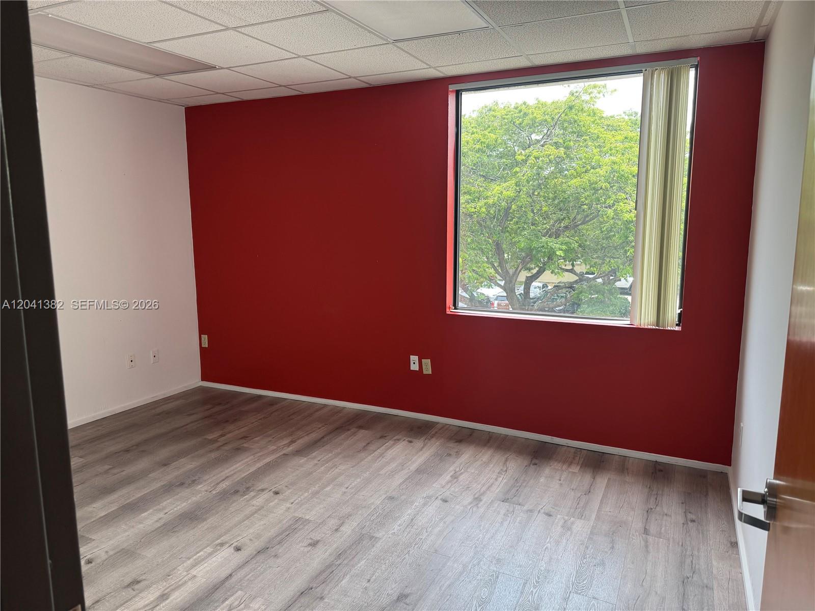 An empty office room features a red accent wall, wood-look flooring, and a large window — 8700 W Flagler St # 200, Miami FL 33174, MIAMI, FL