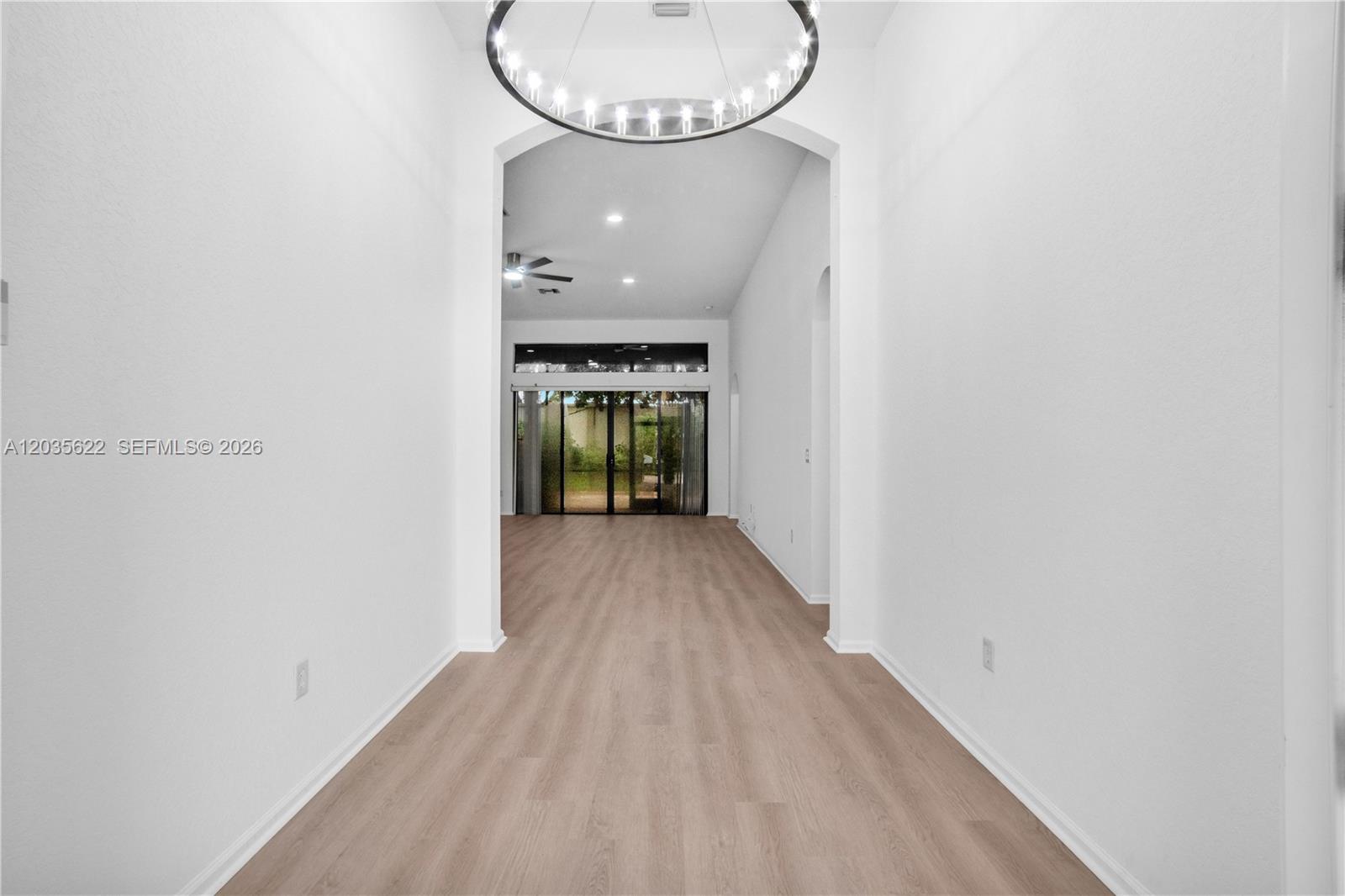 A bright, empty hallway with light wood-look flooring leads to a living area with glass doors — 7747 Via Grande, Boynton Beach FL 33437, BOYNTON BEACH, FL