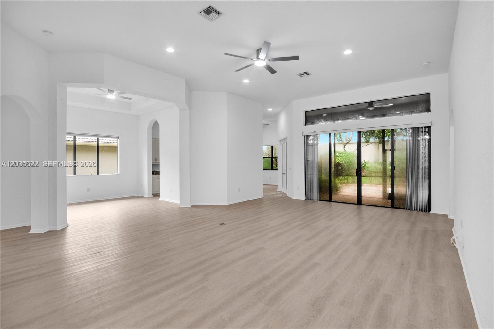 An empty living room features light wood-look flooring, white walls, and large sliding glass doors — 7747 Via Grande, Boynton Beach FL 33437, BOYNTON BEACH, FL