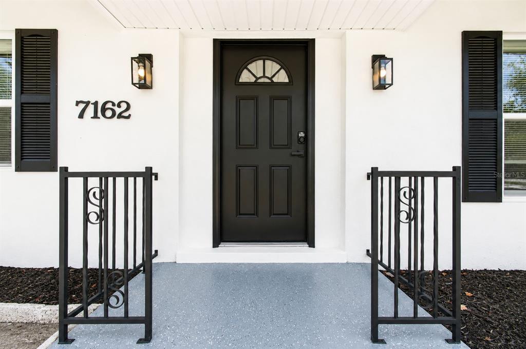 A front entryway features a black door, two wall-mounted lanterns, and black metal railings — 7162 CAMBRIDGE STREET, SPRING HILL, FL