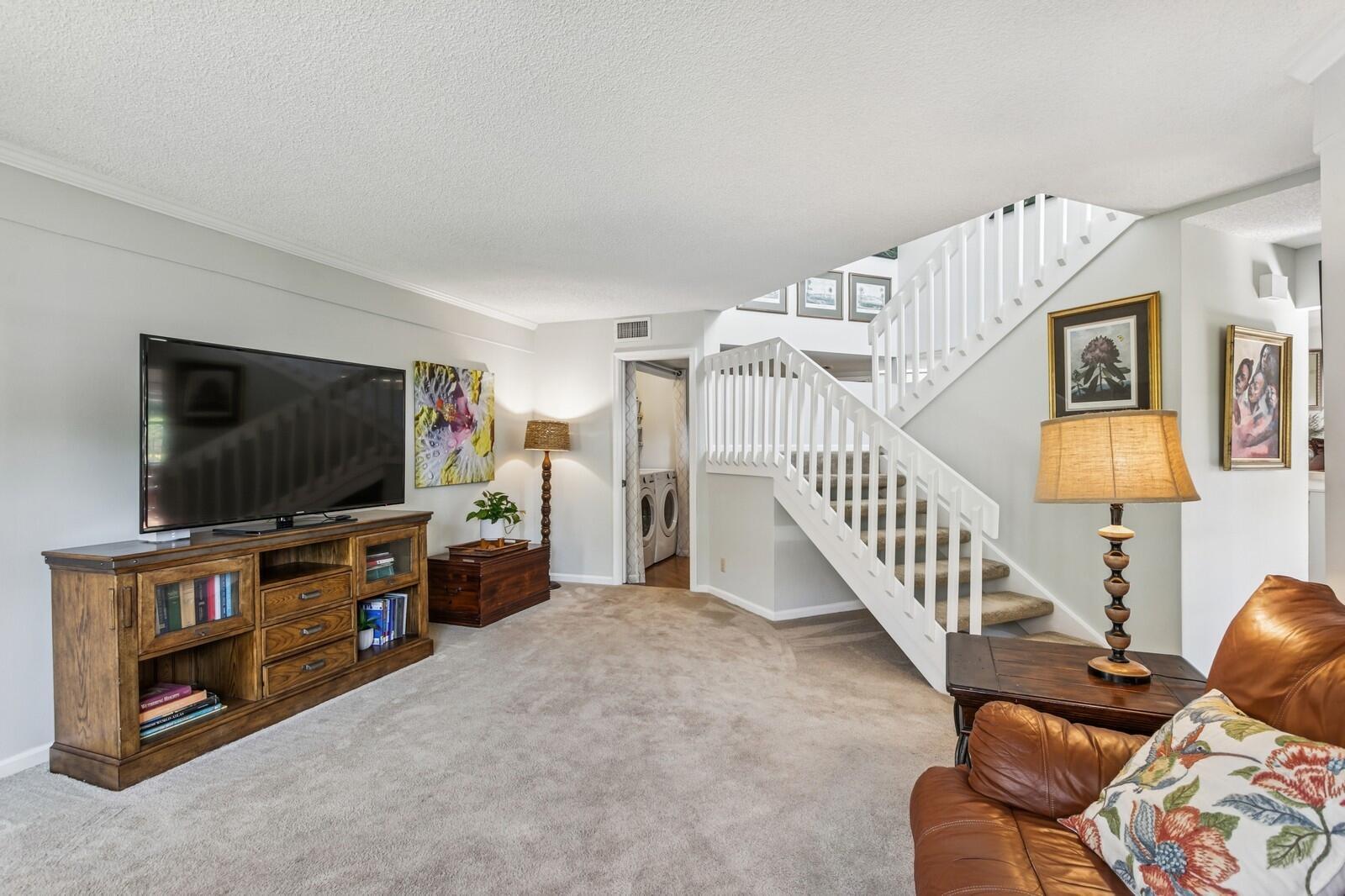 A living room features a television console, a staircase with white railings, and carpeted flooring — 24 Uno Lago Drive, Juno Beach FL 33408, JUNO BEACH, FL