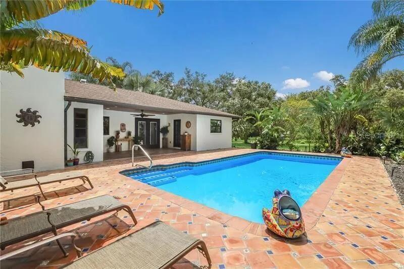A swimming pool sits on a tiled patio next to a white house with a porch — 2077 RACIMO DRIVE, SARASOTA, FL