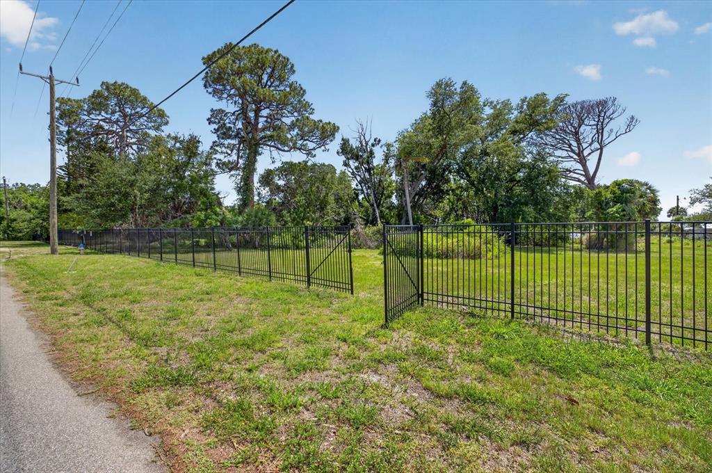 A black metal fence encloses a grassy vacant lot with mature trees under a blue sky — 8258 LONGBAY BLVD, SARASOTA, FL