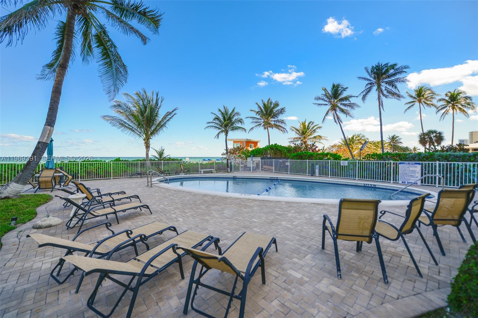 A swimming pool is surrounded by lounge chairs on a paved patio with palm trees — 1936 S Ocean Dr # 2D, Hallandale Beach FL 33009, HALLANDALE BEACH, FL