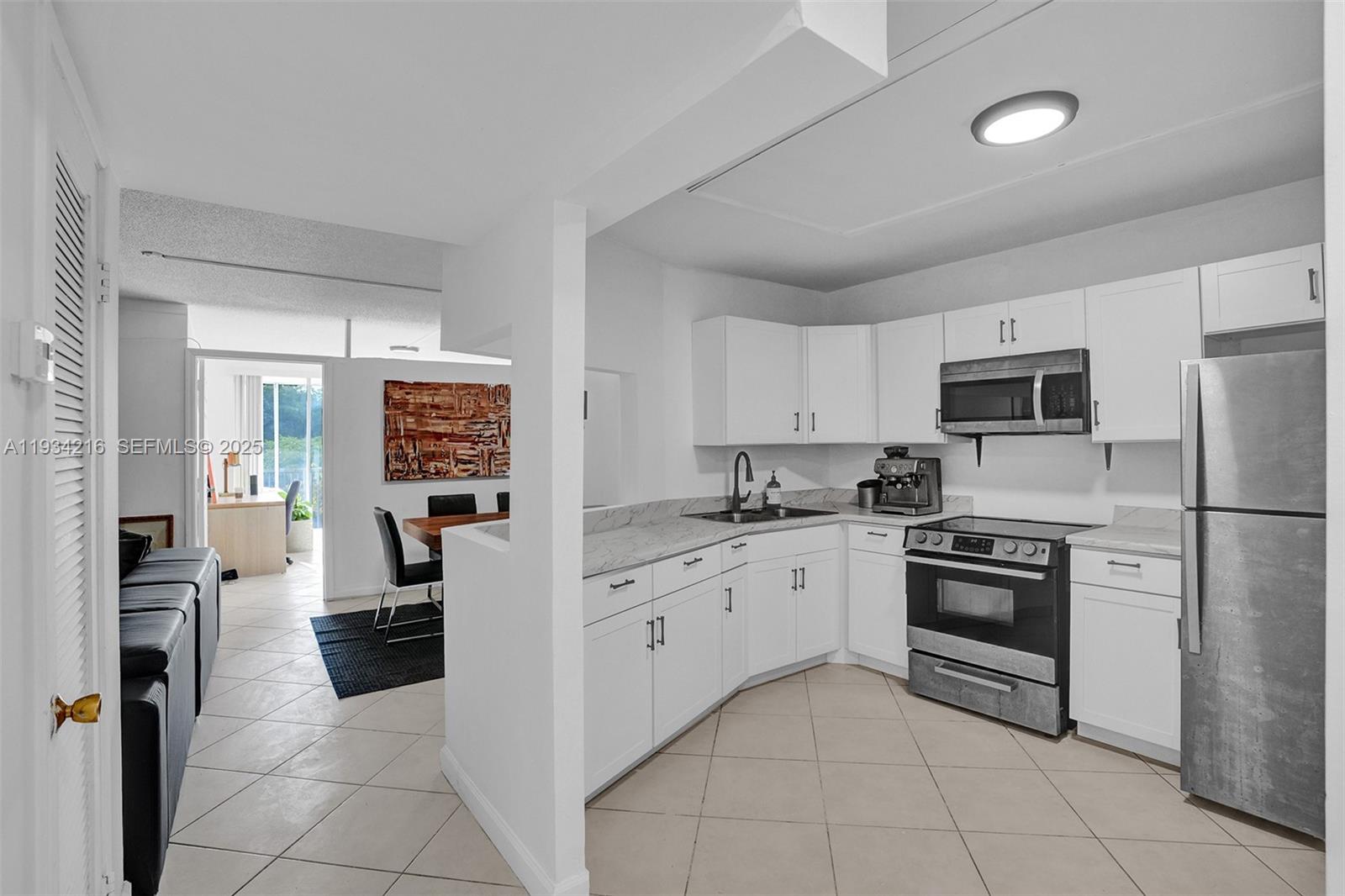 A white kitchen with stainless steel appliances and marble-look countertops opens to a dining area — 1780 NE 191st St # 2, Miami FL 33179, MIAMI, FL