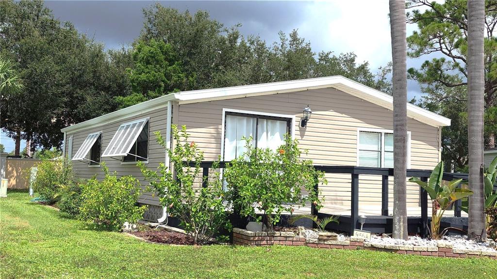 A single-story mobile home with tan siding, a dark wooden deck, and surrounding landscaping — 2100 KINGS HIGHWAY UNIT 956A, PORT CHARLOTTE, FL