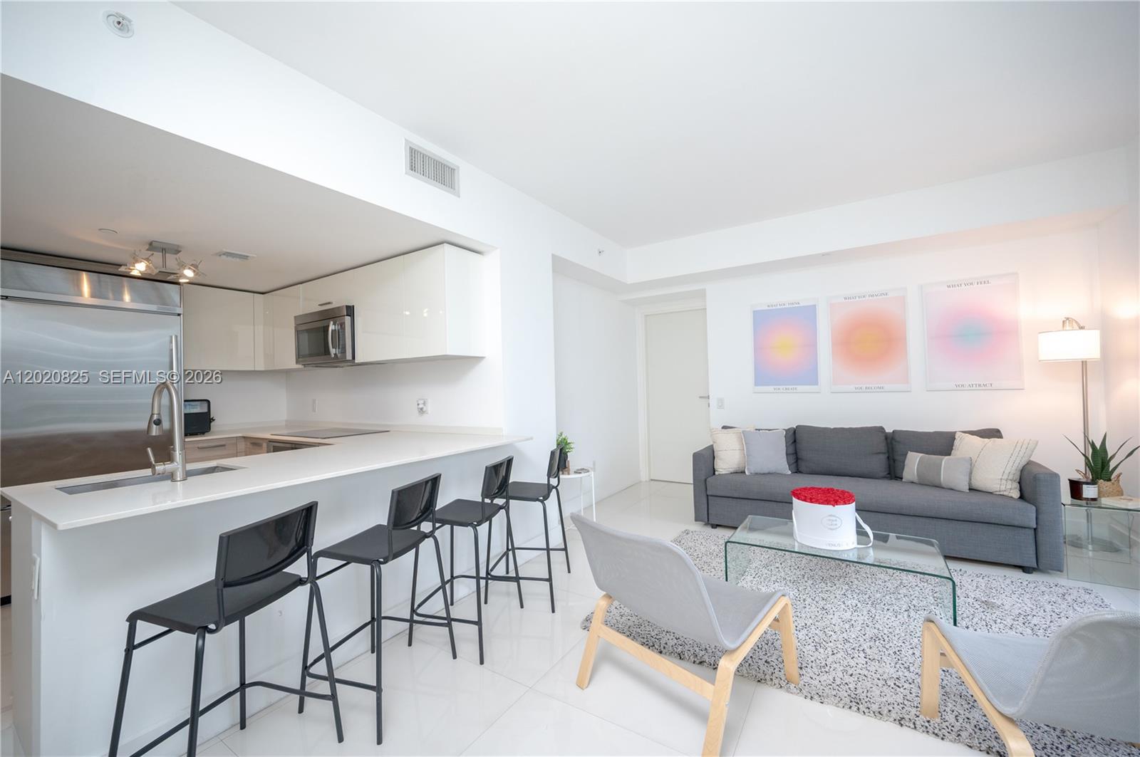 A modern open-concept kitchen and living area features white cabinetry, bar seating, and gray furniture — 1300 Brickell Bay Dr # 1601, Miami FL 33131, MIAMI, FL