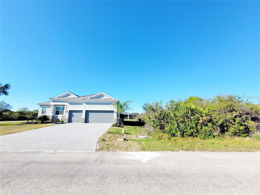 A single-story house with a gray garage and a paved driveway sits beside overgrown vegetation — 14442 AMESTOY AVENUE, PORT CHARLOTTE, FL