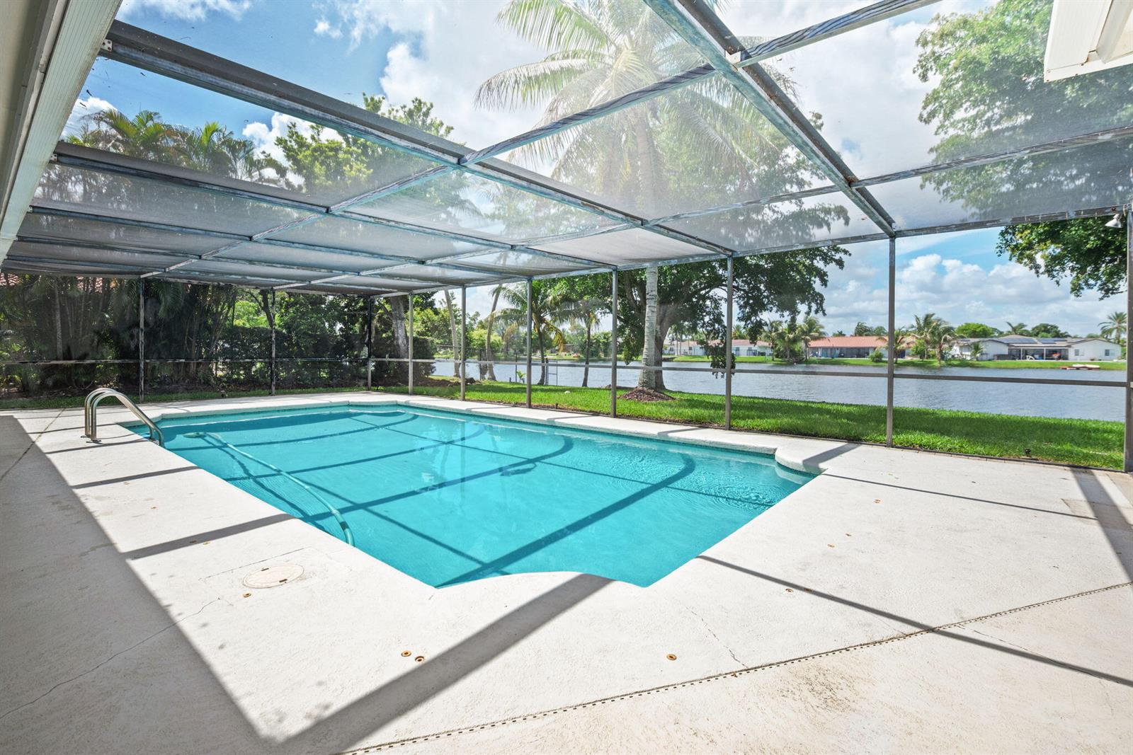 A screened-in swimming pool overlooks a lake with palm trees and houses in the background — 16731 Harbor Court, Weston FL 33326, WESTON, FL