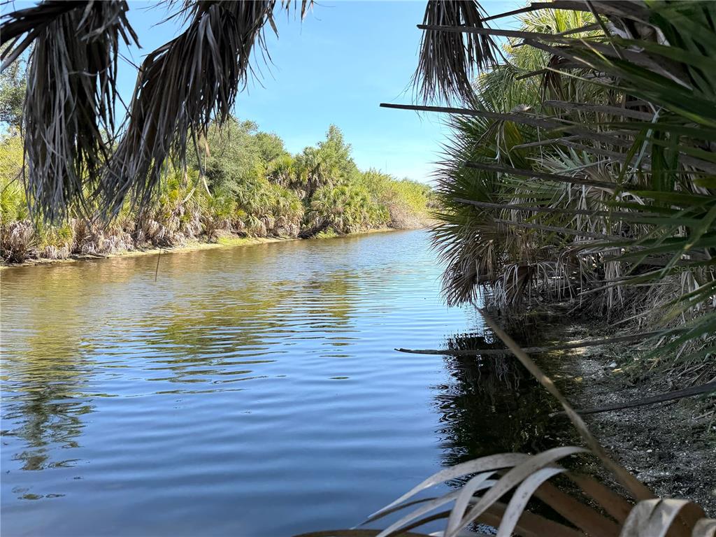 A narrow, calm canal flows through a lush, green landscape with palm trees and vegetation — 7418 SIOUX STREET, PORT CHARLOTTE, FL
