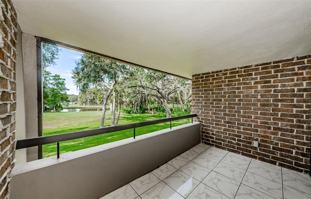 A balcony with white marble-look tile flooring, a brick wall, and a view of trees — 36750 US HIGHWAY 19 N UNIT 2177, PALM HARBOR, FL