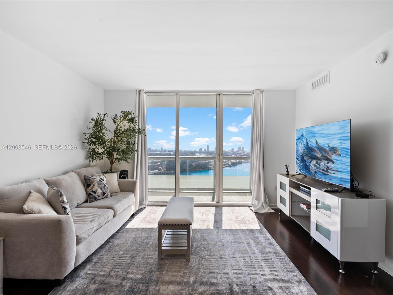 A living room features a light gray sofa, a media console, and floor-to-ceiling windows — 50 Biscayne Blvd # 3308, Miami FL 33132, MIAMI, FL