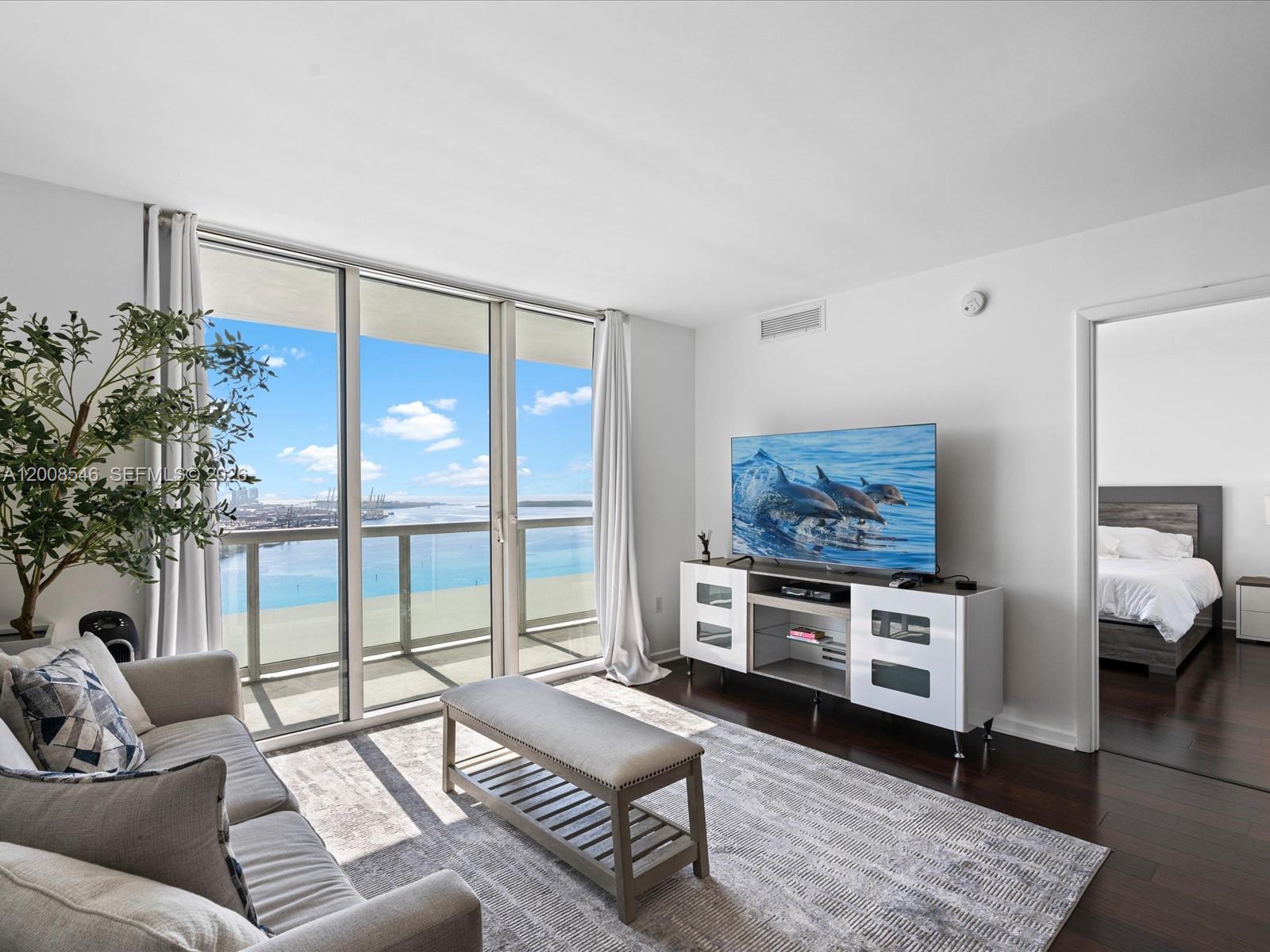 A living room features a sofa, television console, and floor-to-ceiling windows overlooking the ocean — 50 Biscayne Blvd # 3308, Miami FL 33132, MIAMI, FL