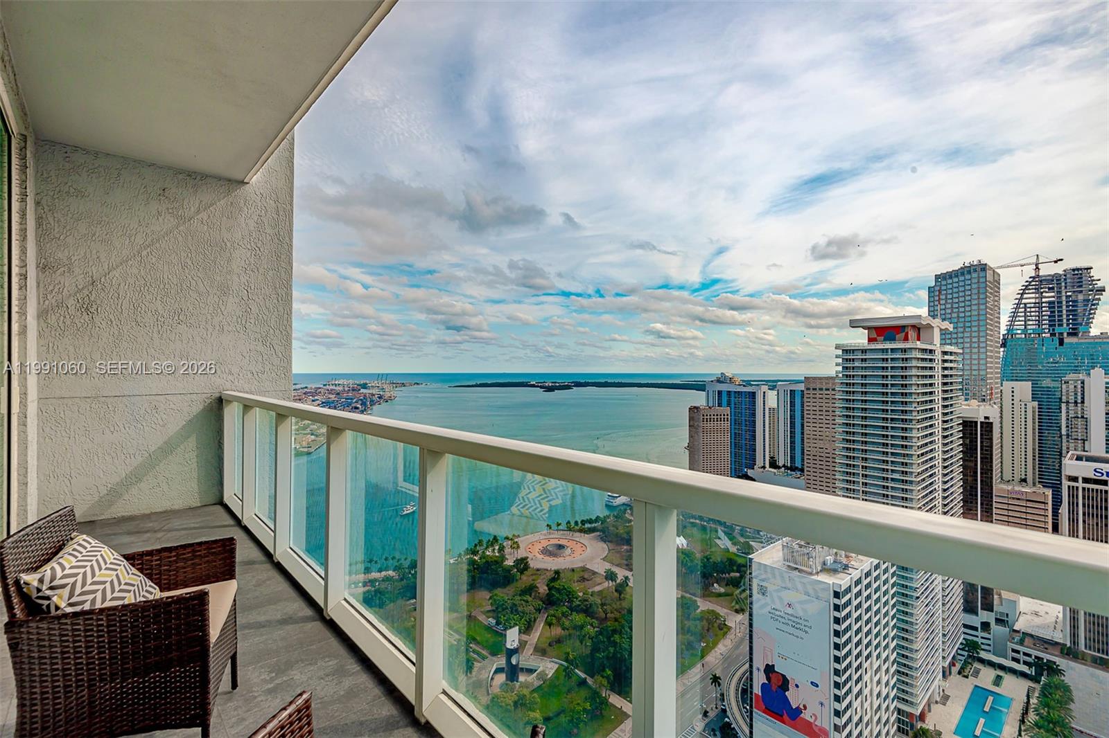 A balcony with wicker furniture overlooks a city skyline, a park, and the ocean — 244 Biscayne Blvd # 4907, Miami FL 33132, MIAMI, FL