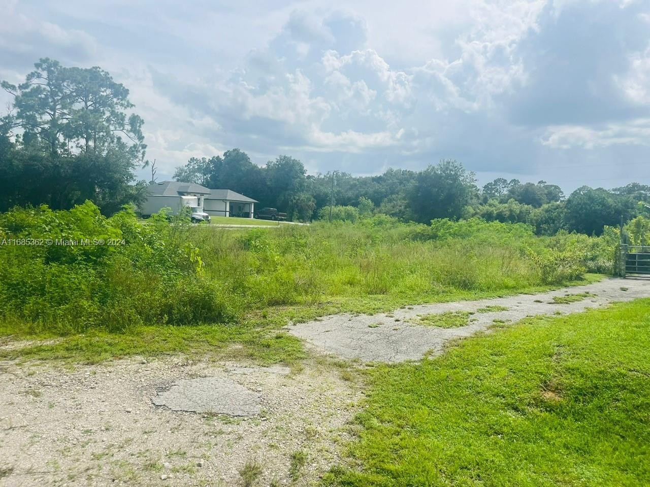 An overgrown, grassy vacant lot features a gravel driveway and a neighboring house in the background — 805 E 6 st, Lehigh Acres FL 33972, LEHIGH ACRES, FL
