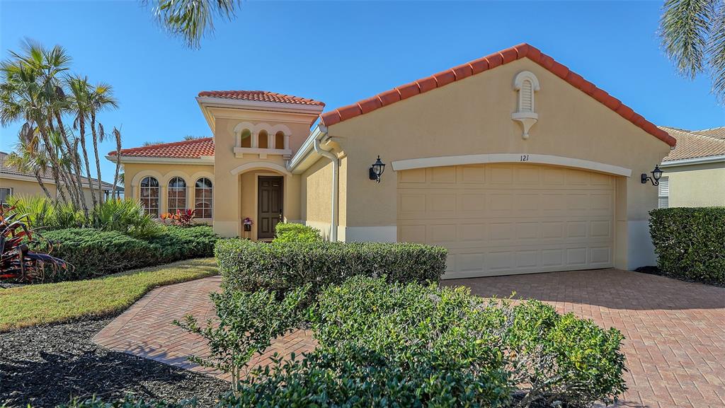 A tan stucco house features a red tile roof, a two-car garage, and landscaped shrubs — 121 SAVONA WAY, NOKOMIS, FL