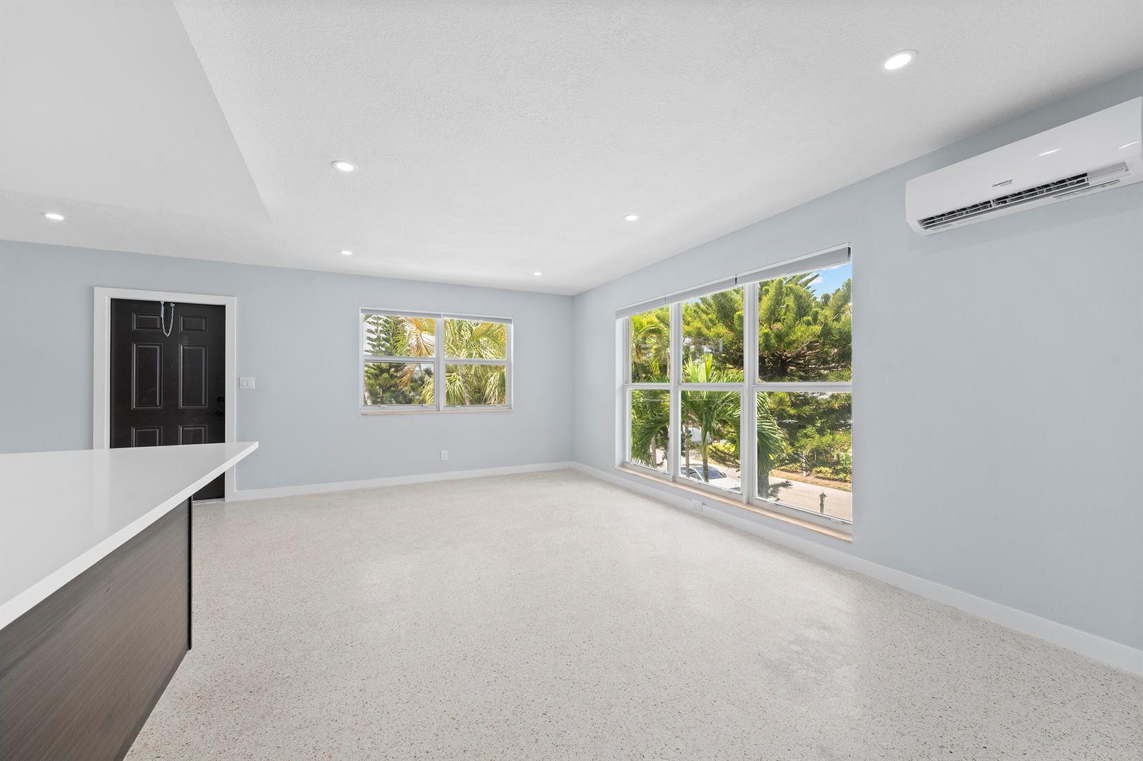 A bright, empty room features light blue walls, terrazzo flooring, large windows, and a wall-mounted air conditioner — 3220 NE 7th # 2, Pompano Beach FL 33062, POMPANO BEACH, FL