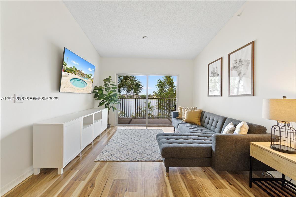 A living room features a grey sectional sofa, a white media console, and a balcony view — 5065 Wiles Rd # 308, Coconut Creek FL 33073, COCONUT CREEK, FL