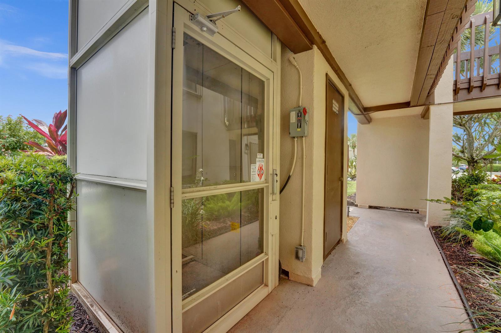An outdoor walkway features a glass-enclosed elevator shaft next to a small utility door — 5306 Cedar Lake Drive # 201, Boynton Beach FL 33437, BOYNTON BEACH, FL