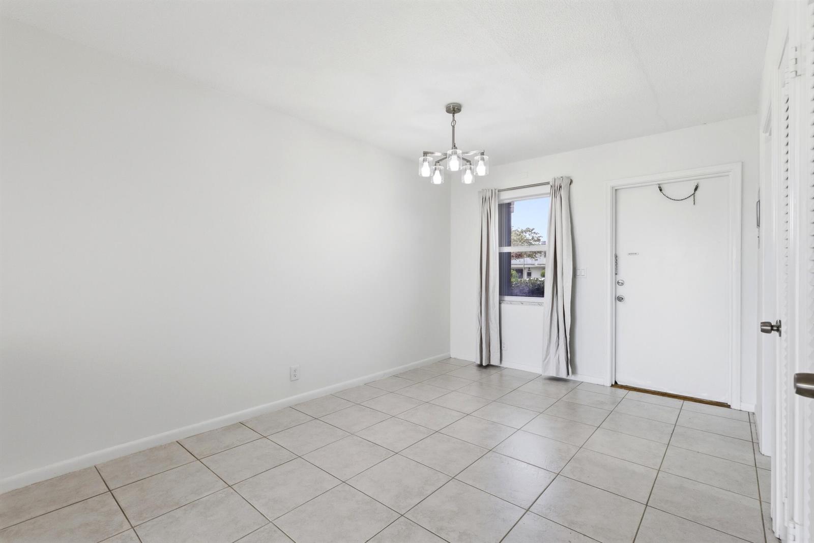 An empty room features white walls, light-colored tile flooring, a chandelier, and a white door — 311 E Ocean Avenue # 104, Lantana FL 33462, LANTANA, FL