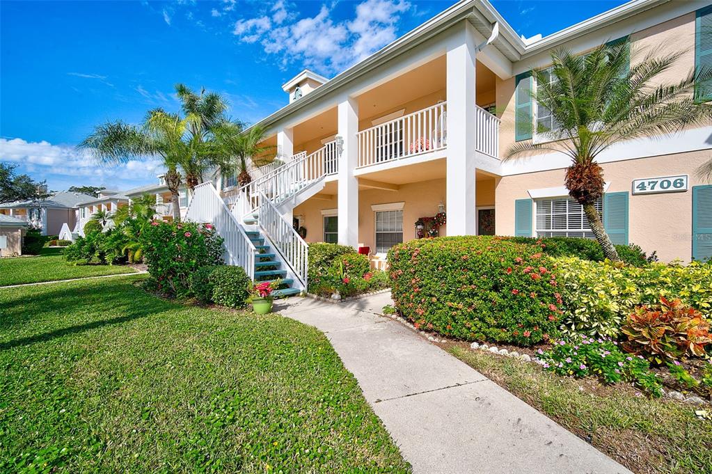 A two-story apartment building exterior features a white staircase, balconies, and landscaped front gardens — 4706 SAND TRAP STREET CIRCLE E UNIT 202, BRADENTON, FL