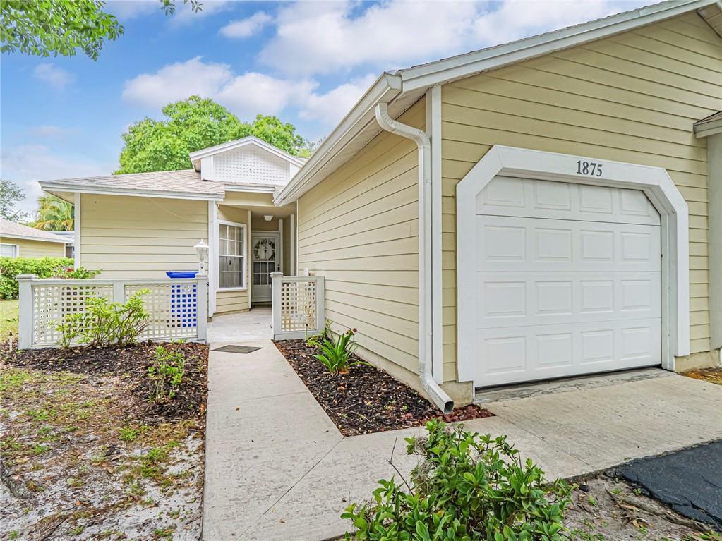 A beige house exterior features a white garage door, a walkway, and a small fence — 1875 Sixty Oaks Lane, Vero Beach FL 32966, VERO BEACH, FL
