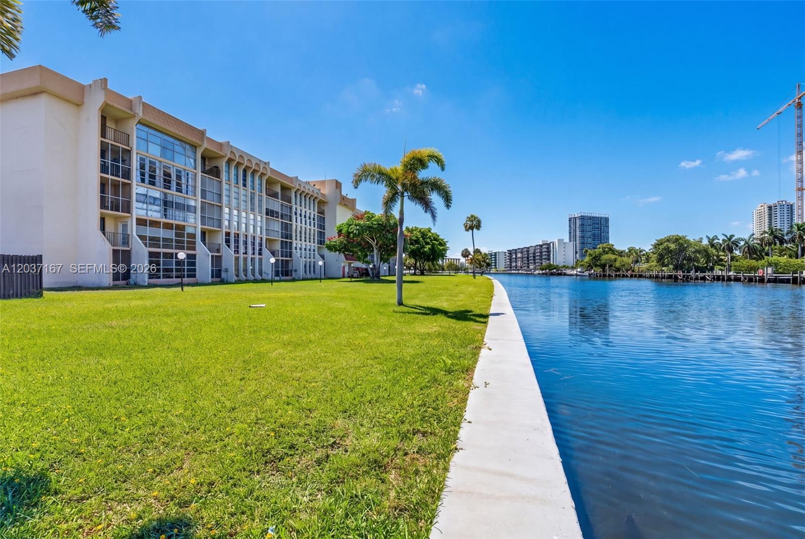 A multi-story apartment building sits beside a canal with a grassy lawn and palm trees — 601 Three Islands Blvd # 403, Hallandale Beach FL 33009, HALLANDALE BEACH, FL