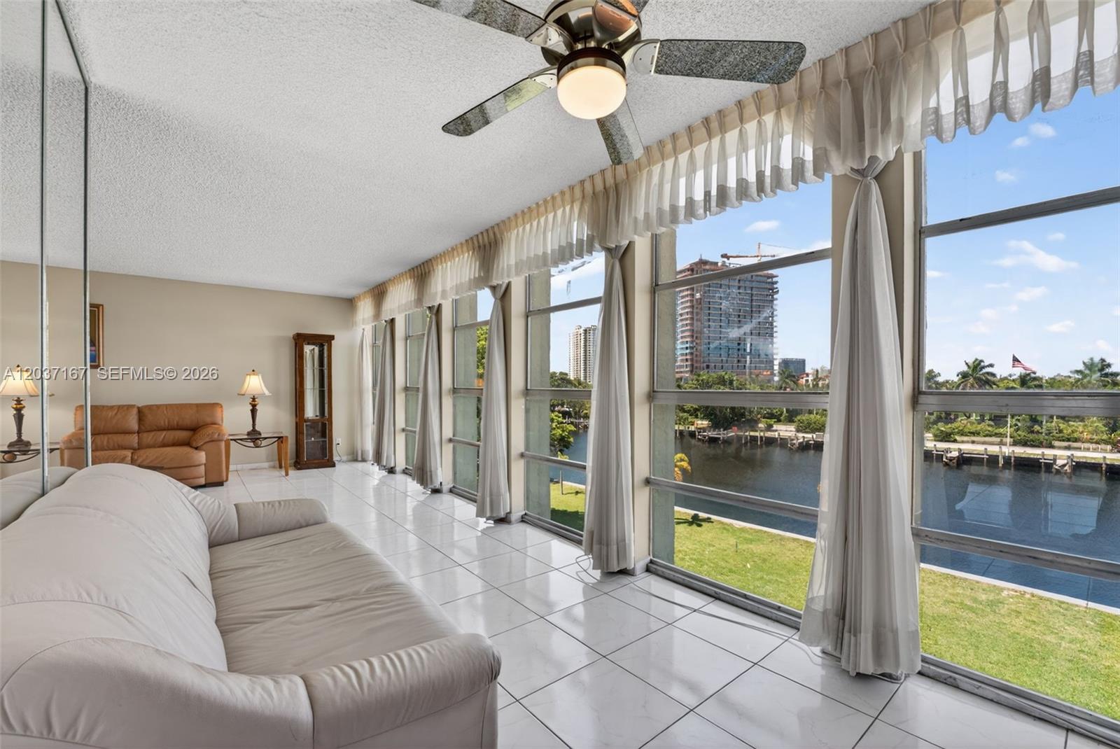 A living room with white tile floors, two sofas, and floor-to-ceiling windows overlooking a waterway — 601 Three Islands Blvd # 403, Hallandale Beach FL 33009, HALLANDALE BEACH, FL