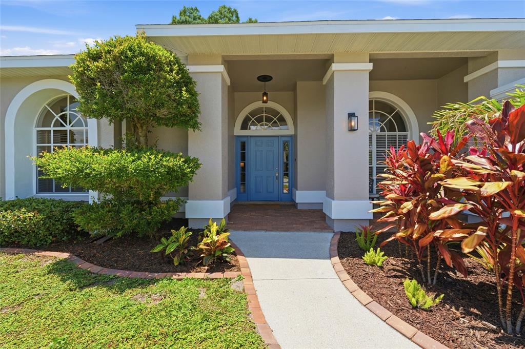 A concrete walkway leads to the blue front door of a beige house with arched windows — 6130 55TH AVENUE CIRCLE E, BRADENTON, FL