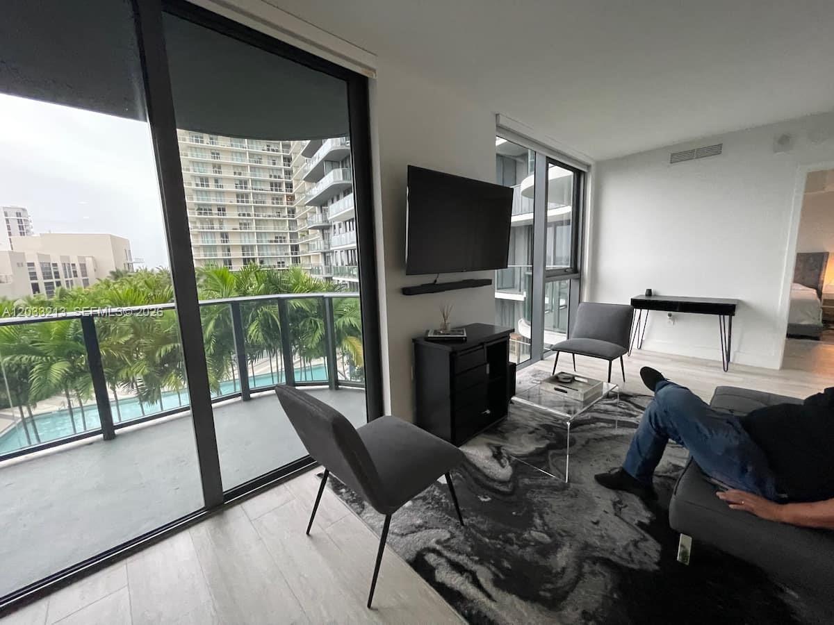 A living room features a television, gray furniture, a patterned rug, and floor-to-ceiling windows — 121 NE 34th St # 1015, Miami FL 33137, MIAMI, FL