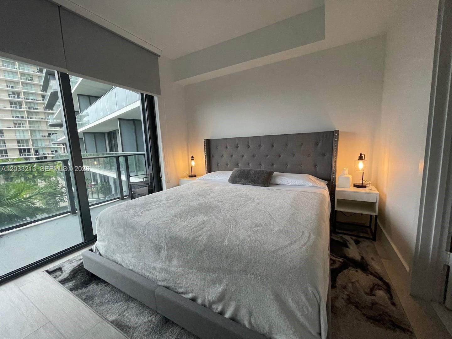 A bedroom features a tufted gray headboard, two bedside lamps, and a balcony view — 121 NE 34th St # 1015, Miami FL 33137, MIAMI, FL