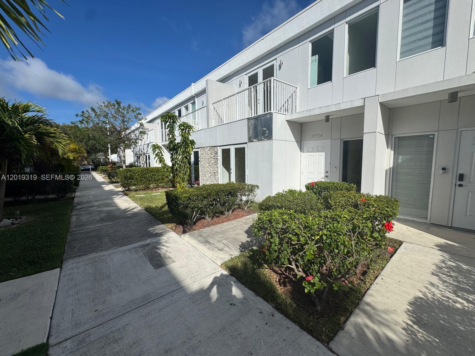 A modern white townhouse exterior features a small balcony, a front door, and landscaped bushes — 13724 SW 259th St, Homestead FL 33032, HOMESTEAD, FL
