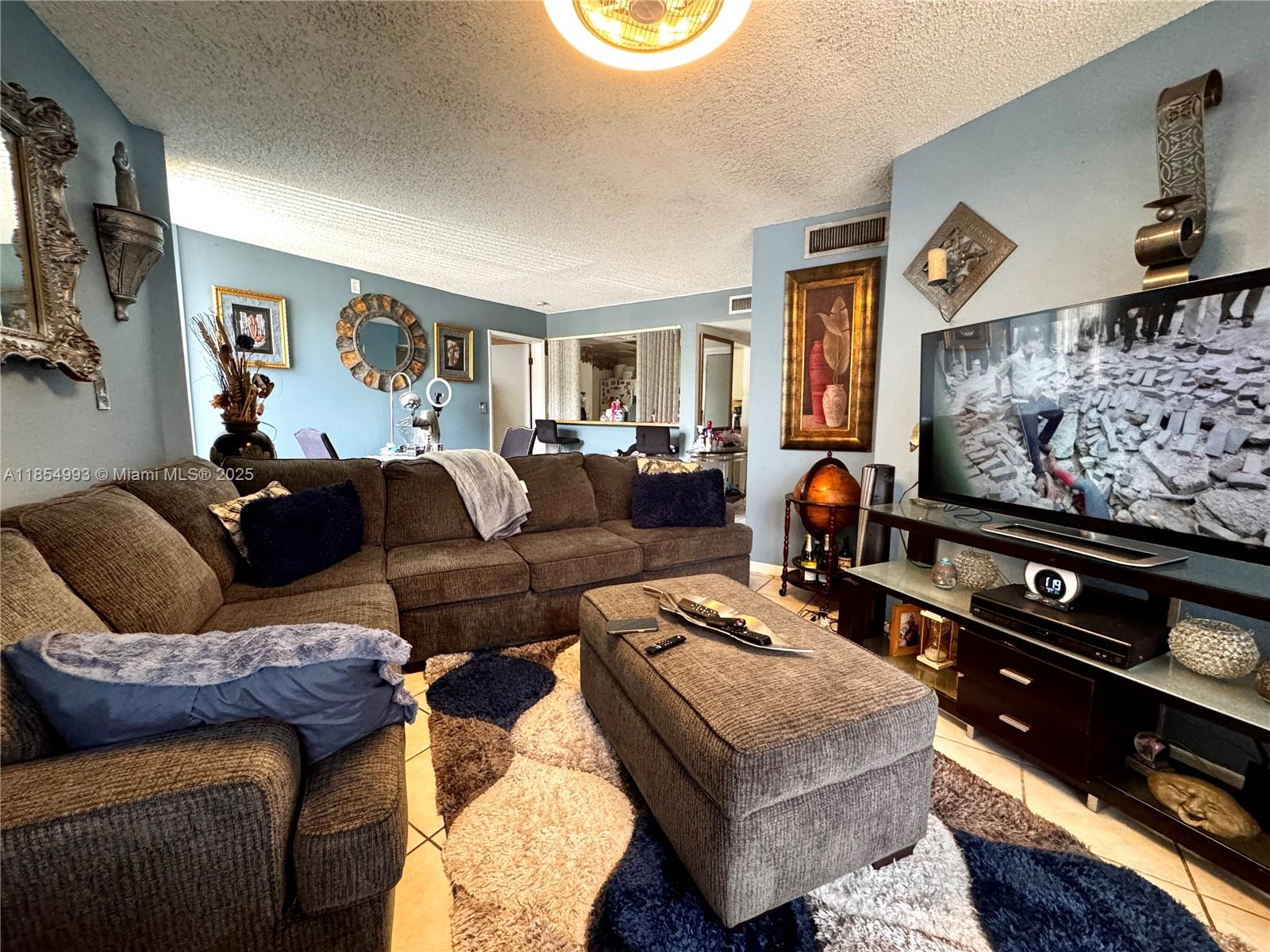 A living room features a large brown sectional sofa, an ottoman, and a wall-mounted television — 7165 NW 186th St # A211, Hialeah FL 33015, HIALEAH, FL