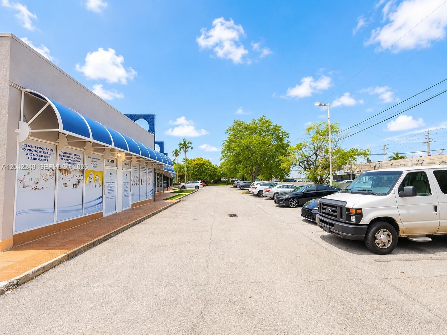 A commercial building with blue awnings stands beside a parking lot with several cars — 10240 SW 56th St # C, Miami FL 33165, MIAMI, FL