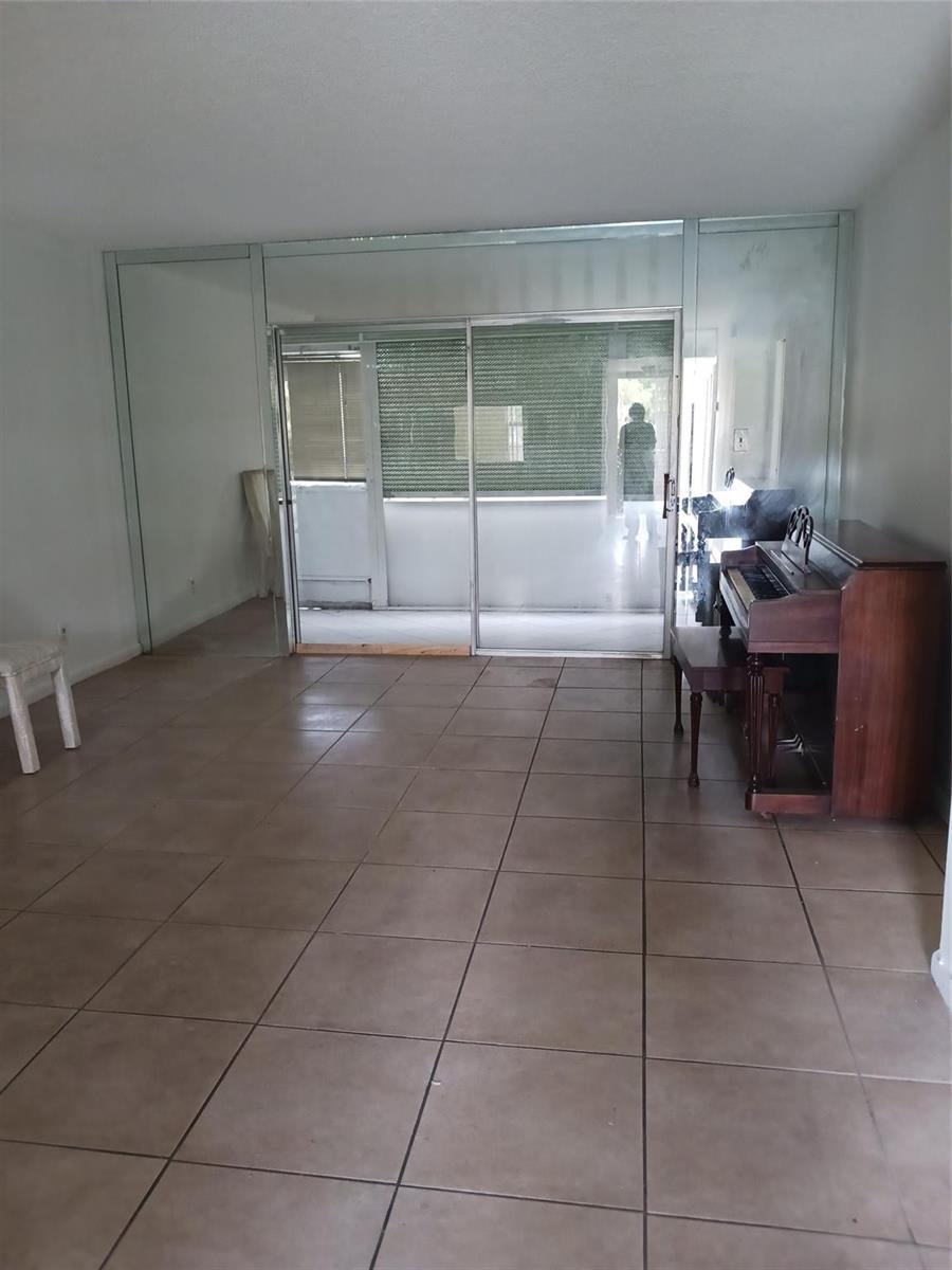 An empty room with tiled floors, a piano, and sliding glass doors leading to a balcony — 7605 W Atlantic Boulevard # 106, Margate FL 33063, MARGATE, FL
