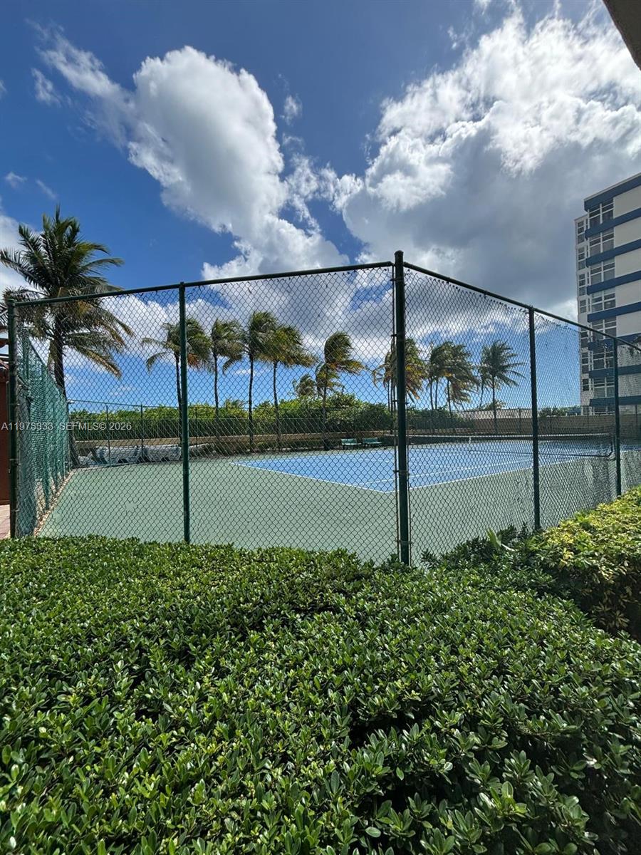 A fenced tennis court is surrounded by palm trees and a hedge under a blue sky — 2080 S Ocean Dr # 308, Hallandale Beach FL 33009, HALLANDALE BEACH, FL