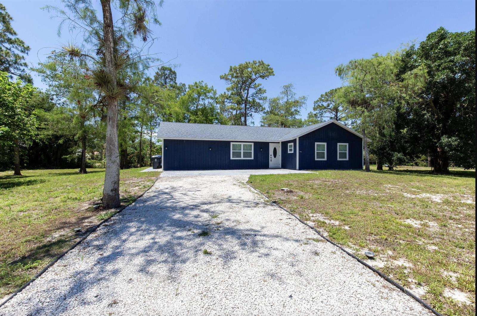 A blue single-story house with a white door and a gravel driveway sits among trees — 16932 W Aintree Drive W, Loxahatchee FL 33470, LOXAHATCHEE, FL