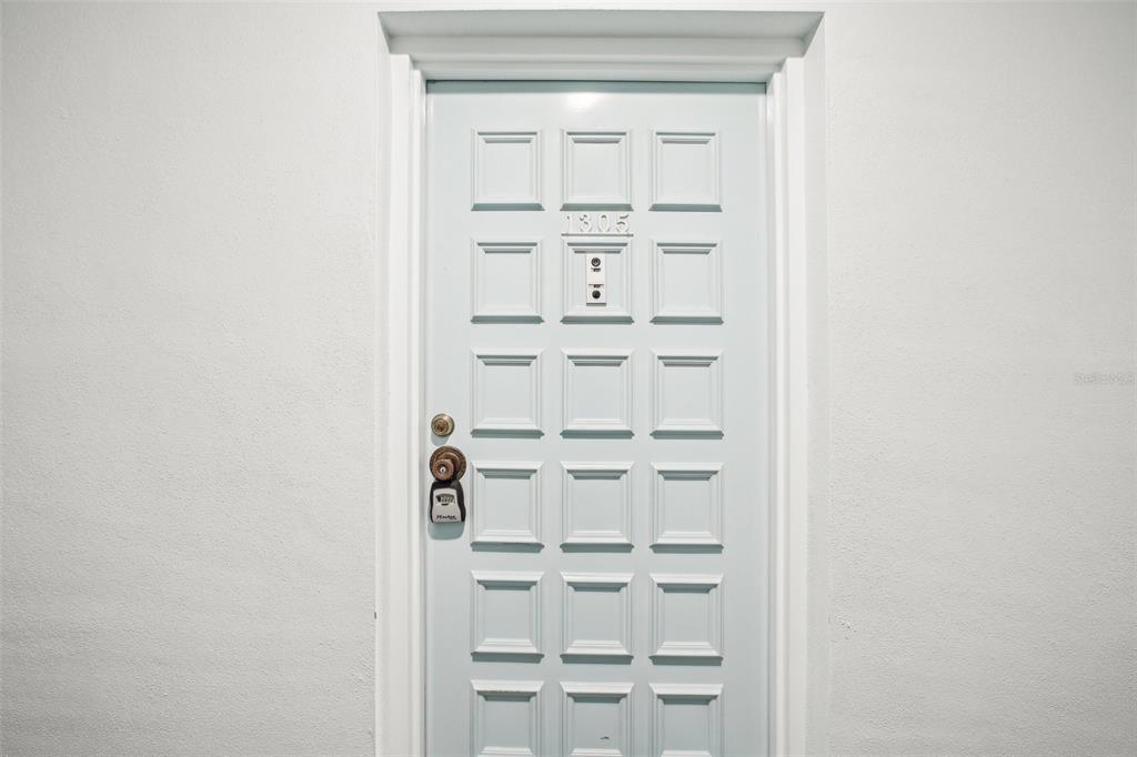 A white paneled front door with a lockbox and a peephole is set in a wall — 2700 BAYSHORE BOULEVARD UNIT 1305, DUNEDIN, FL