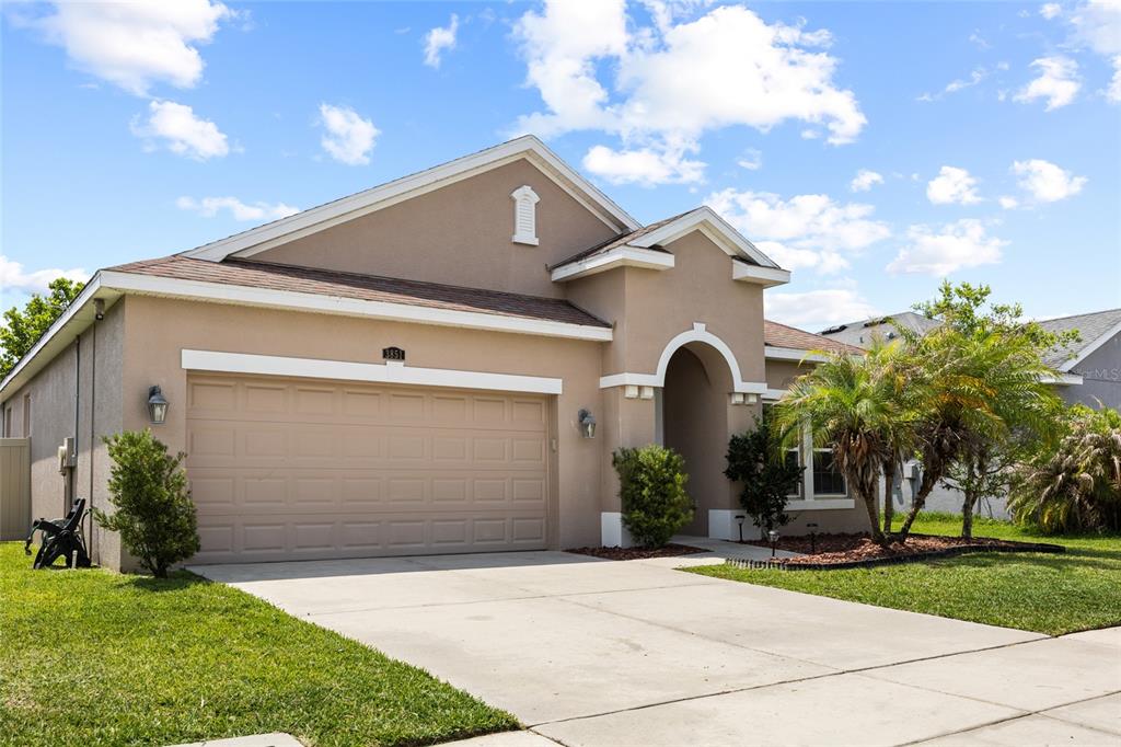 A single-story tan stucco house features a two-car garage and a covered entryway — 3851 PACENTE LOOP, WESLEY CHAPEL, FL