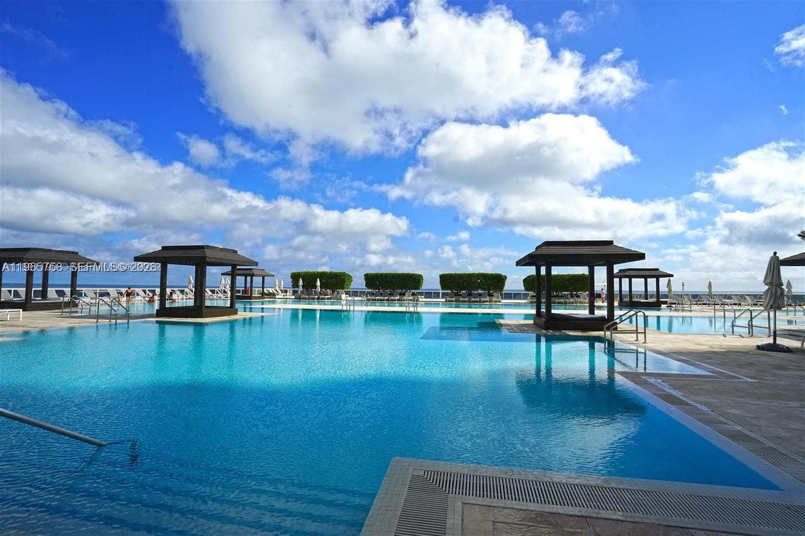 A large outdoor swimming pool features several wooden cabanas under a bright, cloudy sky — 1800 S Ocean Dr # 2406, Hallandale Beach FL 33009, HALLANDALE BEACH, FL