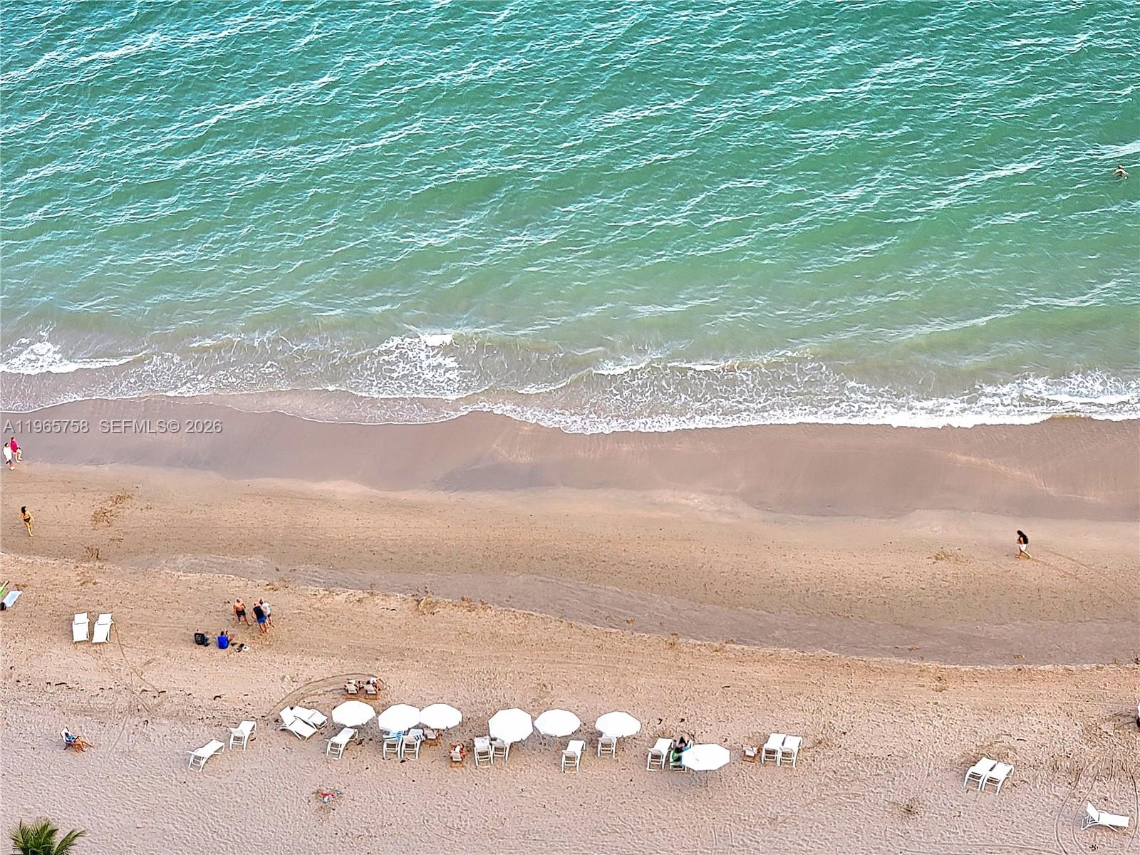 An aerial view shows a sandy beach with lounge chairs, umbrellas, and people near water — 1800 S Ocean Dr # 2406, Hallandale Beach FL 33009, HALLANDALE BEACH, FL