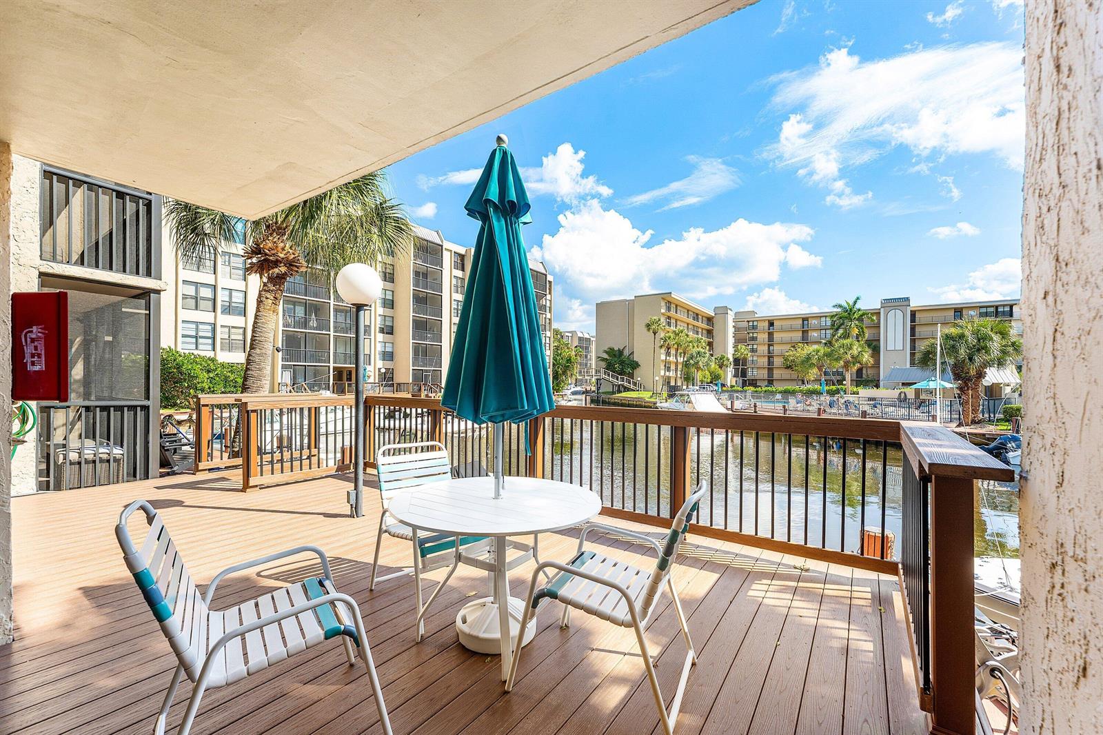 A wooden deck features a patio table with a teal umbrella overlooking a canal — 10 Royal Palm Way # 203, Boca Raton FL 33432, BOCA RATON, FL