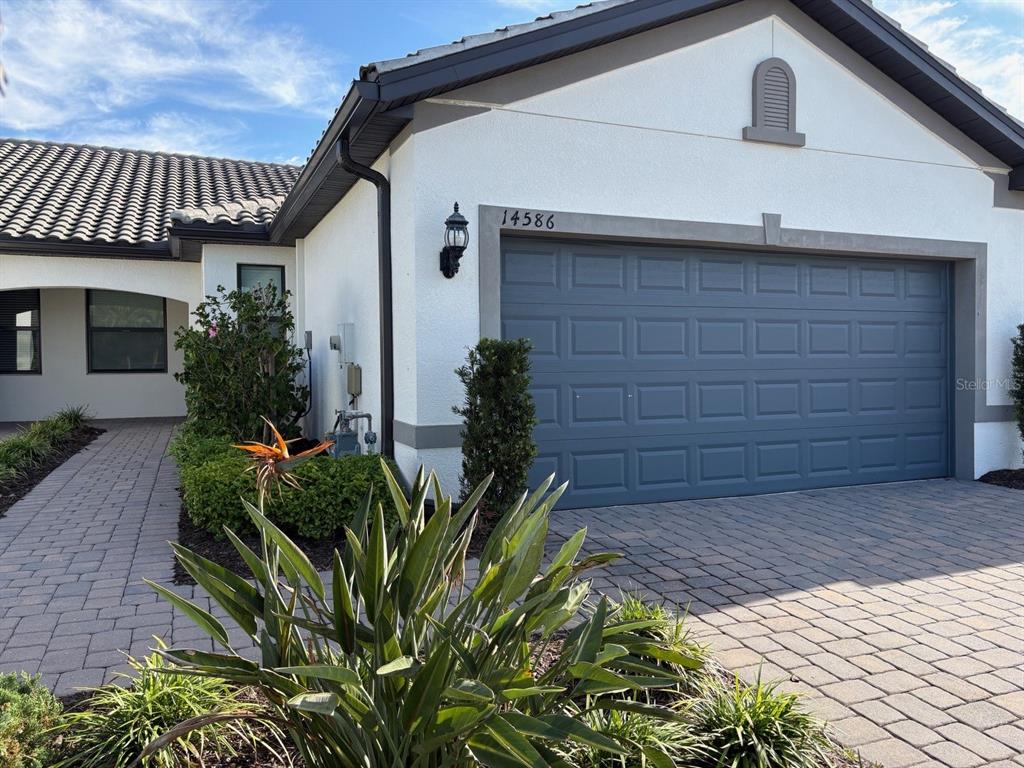 A single-story house exterior features a gray garage door, white stucco walls, and paver driveway — 14586 MOSSY PINE COURT, NOKOMIS, FL