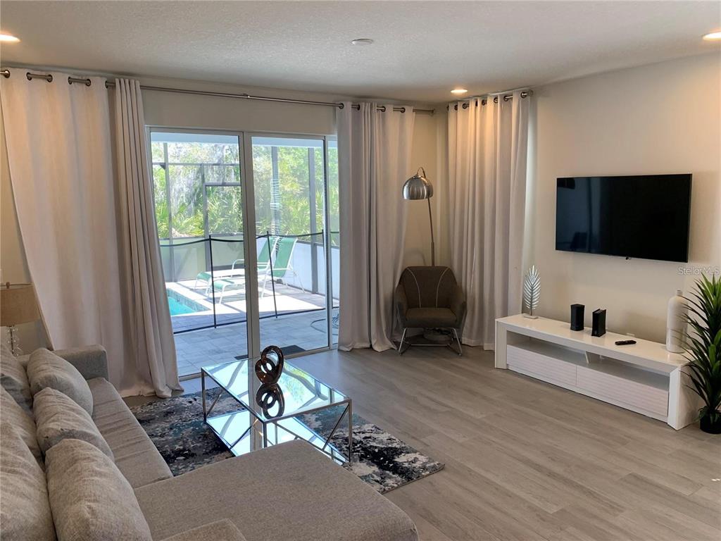 A living room features a gray sectional, glass coffee table, media console, and sliding glass doors — 17307 HIDDEN FOREST DRIVE, CLERMONT, FL